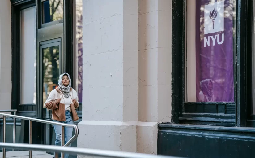 Jovem usando casaco e cachecol do lado de fora de um prédio com banner da NYU, em ambiente urbano, sugerindo intercâmbio nos EUA.